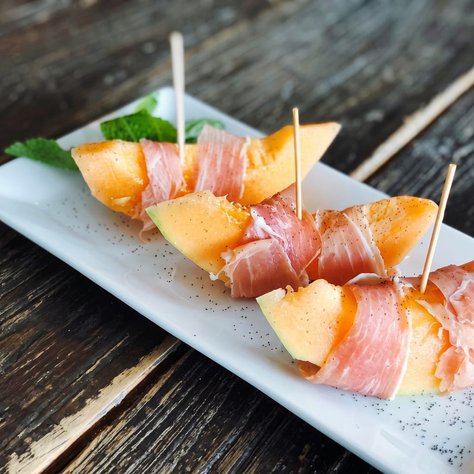 Prosciutto-wrapped melon skewers arranged on a white plate with greens and black sesame seeds.