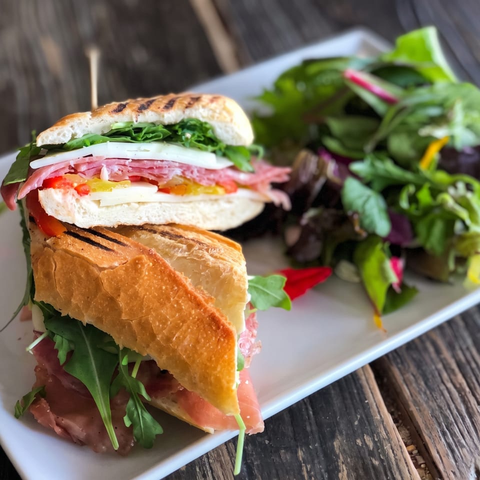 Grilled baguette sandwich layered with greens, sliced meat, cheese, and vegetables, served with a fresh salad on a white plate.